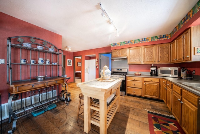 kitchen with stainless steel appliances, brown cabinetry, dark wood finished floors, and under cabinet range hood