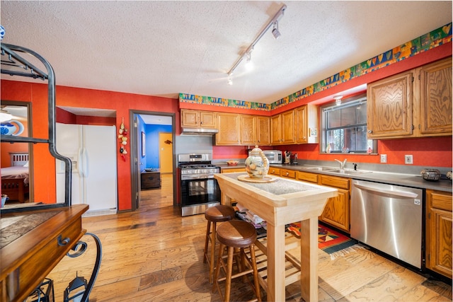 kitchen with brown cabinets, stainless steel appliances, light countertops, under cabinet range hood, and a sink