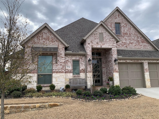 french provincial home with driveway, brick siding, stone siding, and roof with shingles