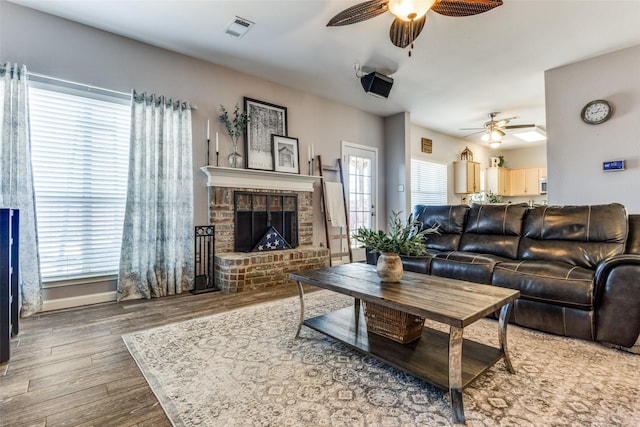 living area with a brick fireplace, a ceiling fan, visible vents, and wood finished floors