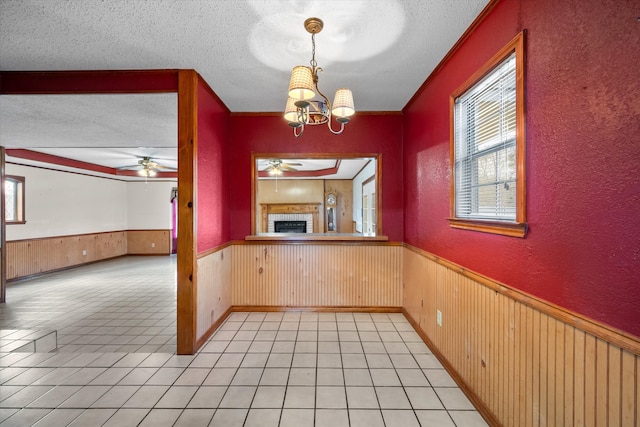 unfurnished dining area with a fireplace, wainscoting, wooden walls, a textured ceiling, and ceiling fan with notable chandelier