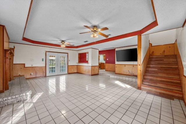 unfurnished living room with light tile patterned floors, wooden walls, french doors, wainscoting, and a tray ceiling