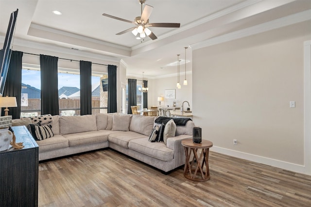 living area with baseboards, wood finished floors, a tray ceiling, crown molding, and ceiling fan with notable chandelier