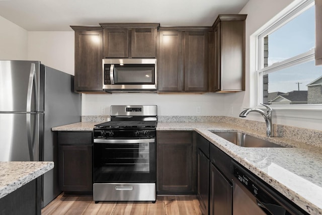 kitchen with dark brown cabinetry, light stone countertops, stainless steel appliances, light wood-type flooring, and a sink