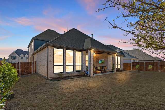 back of house at dusk featuring a patio, a fenced backyard, brick siding, a yard, and roof with shingles