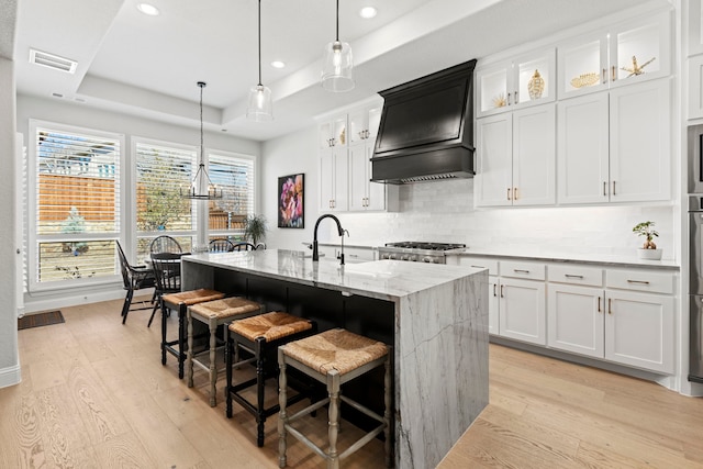 kitchen featuring an island with sink, light wood finished floors, custom range hood, and a raised ceiling