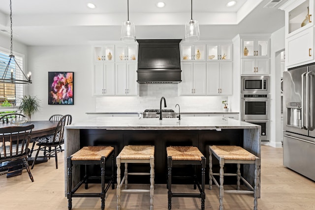 kitchen featuring custom exhaust hood, decorative backsplash, appliances with stainless steel finishes, an island with sink, and light stone countertops