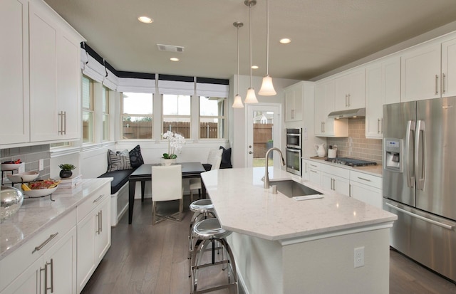 kitchen featuring a kitchen island with sink, under cabinet range hood, a sink, appliances with stainless steel finishes, and decorative light fixtures