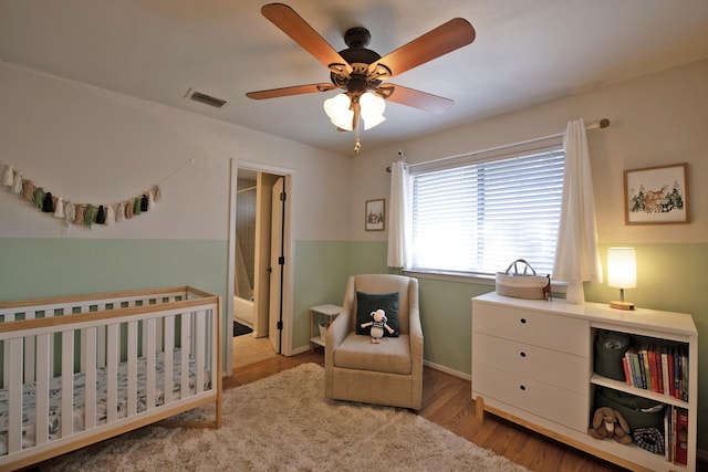 bedroom featuring visible vents, baseboards, a nursery area, ceiling fan, and wood finished floors