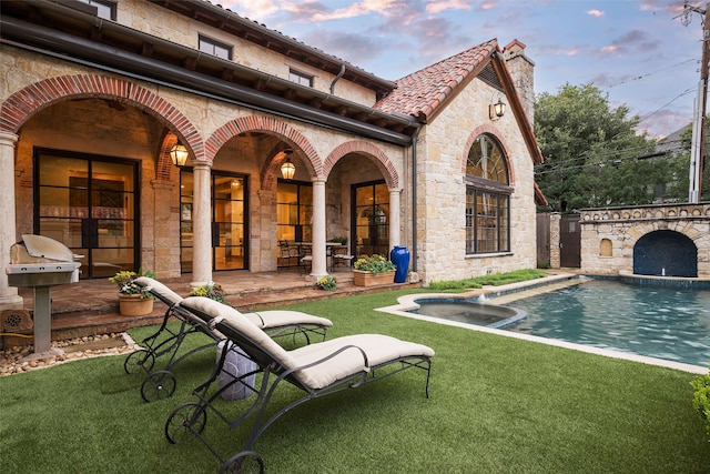 back of property at dusk with a tiled roof, stone siding, an outdoor pool, a chimney, and a patio area