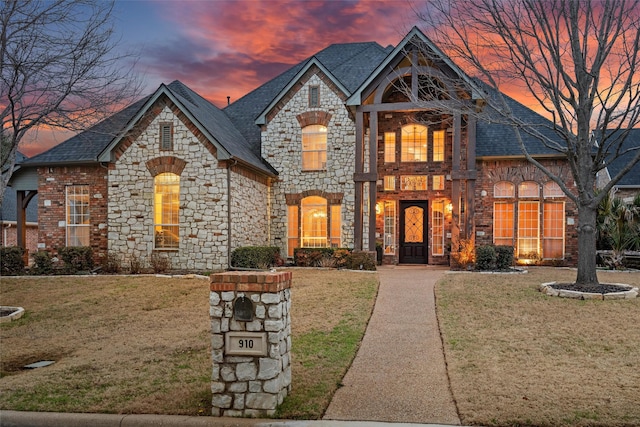 french country home with a shingled roof, stone siding, and a front lawn