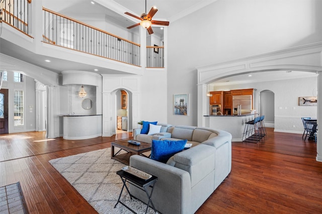 living room featuring baseboards, arched walkways, ceiling fan, ornamental molding, and dark wood-type flooring