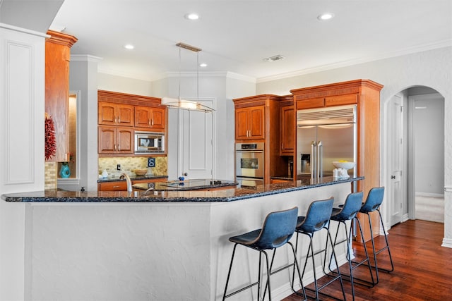 kitchen featuring arched walkways, brown cabinets, decorative light fixtures, a peninsula, and built in appliances