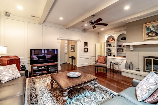 living room featuring a ceiling fan, dark wood-type flooring, beamed ceiling, a fireplace, and a decorative wall