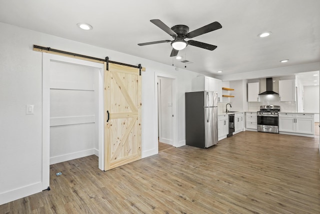 kitchen with open shelves, light wood-style flooring, a barn door, appliances with stainless steel finishes, and wall chimney exhaust hood