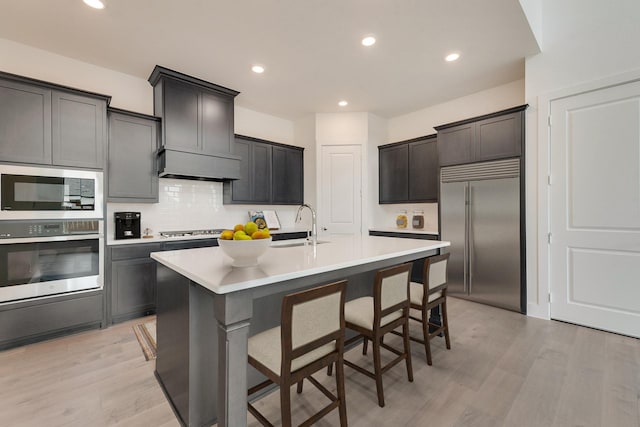 kitchen with light countertops, a center island with sink, light wood finished floors, and built in appliances