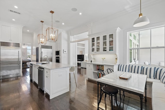 kitchen featuring built in appliances, white cabinetry, hanging light fixtures, an island with sink, and glass insert cabinets