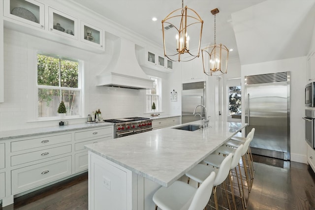 kitchen featuring glass insert cabinets, white cabinets, a sink, built in appliances, and premium range hood