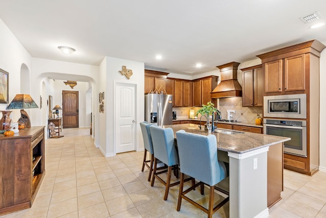 kitchen featuring custom exhaust hood, stainless steel appliances, tasteful backsplash, light stone countertops, and a kitchen bar