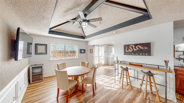 dining area with a textured ceiling, wine cooler, a wainscoted wall, light wood-type flooring, and a raised ceiling