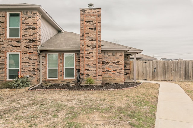 view of property exterior featuring brick siding, roof with shingles, a chimney, a lawn, and fence