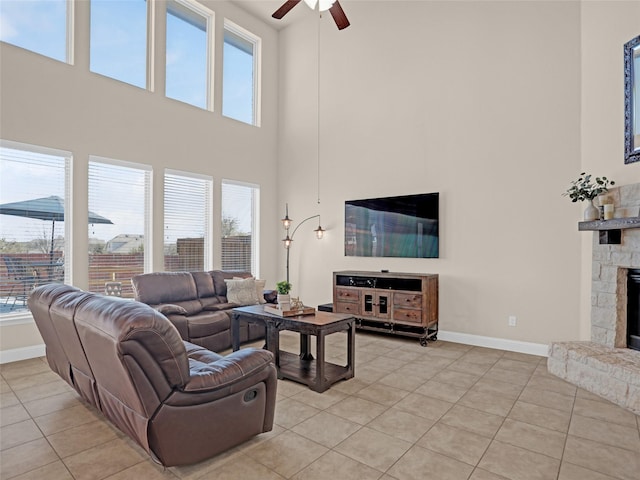 living area with light tile patterned floors, a stone fireplace, a towering ceiling, a ceiling fan, and baseboards