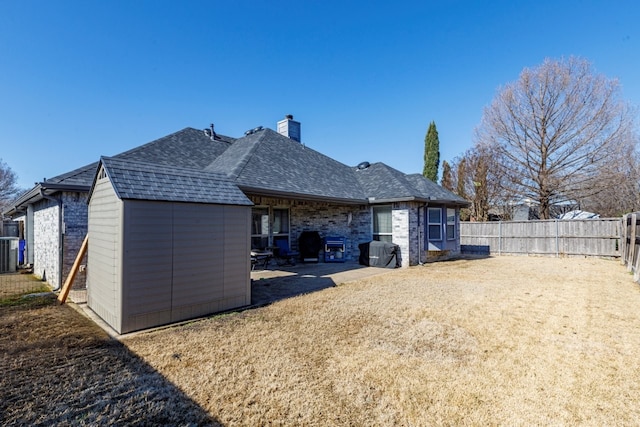 rear view of house featuring brick siding, roof with shingles, a chimney, a fenced backyard, and an outdoor structure