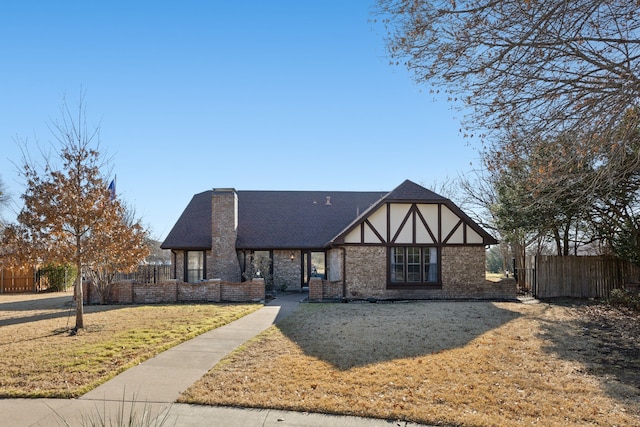 tudor-style house with stucco siding, roof with shingles, fence, a front yard, and brick siding