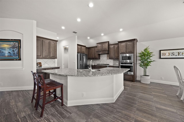 kitchen featuring a center island with sink, appliances with stainless steel finishes, a sink, dark brown cabinets, and light stone countertops