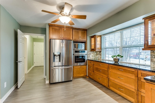 kitchen with tasteful backsplash, appliances with stainless steel finishes, brown cabinetry, glass insert cabinets, and dark stone counters