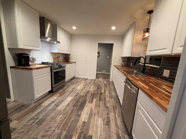 kitchen featuring appliances with stainless steel finishes, white cabinetry, a sink, butcher block countertops, and wall chimney exhaust hood