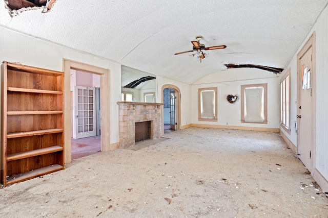 unfurnished living room with a ceiling fan, vaulted ceiling, a stone fireplace, a textured ceiling, and baseboards