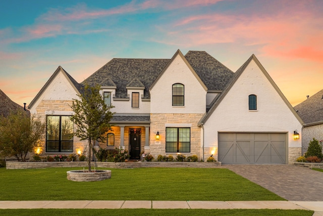 french provincial home featuring roof with shingles, an attached garage, decorative driveway, a front lawn, and brick siding