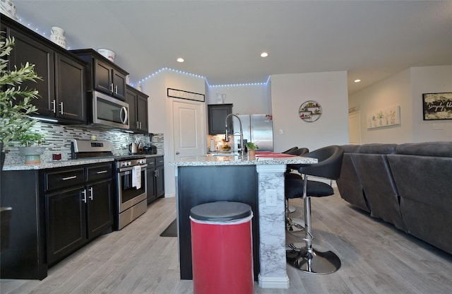 kitchen featuring backsplash, appliances with stainless steel finishes, open floor plan, light wood-type flooring, and a kitchen bar