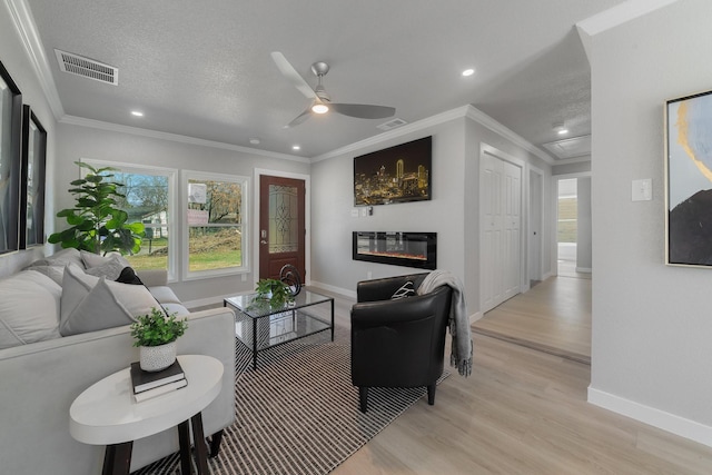 living area with a textured ceiling, visible vents, light wood-style floors, a glass covered fireplace, and crown molding