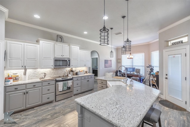 kitchen with arched walkways, an island with sink, appliances with stainless steel finishes, pendant lighting, and a sink