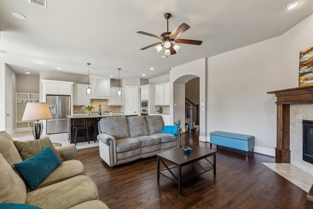 living room featuring baseboards, arched walkways, a ceiling fan, a tile fireplace, and dark wood-type flooring