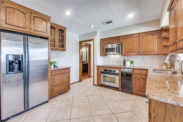 kitchen featuring light stone counters, brown cabinets, stainless steel appliances, glass insert cabinets, and a sink