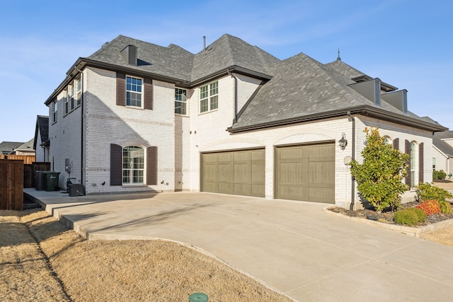 french country style house with a garage, brick siding, fence, concrete driveway, and roof with shingles