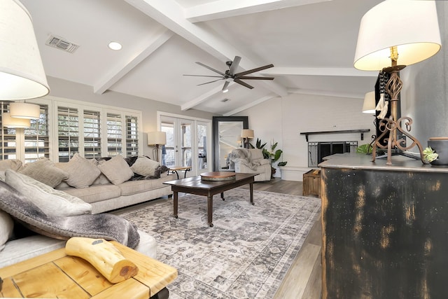 living area featuring vaulted ceiling with beams, visible vents, a fireplace with raised hearth, ceiling fan, and wood finished floors