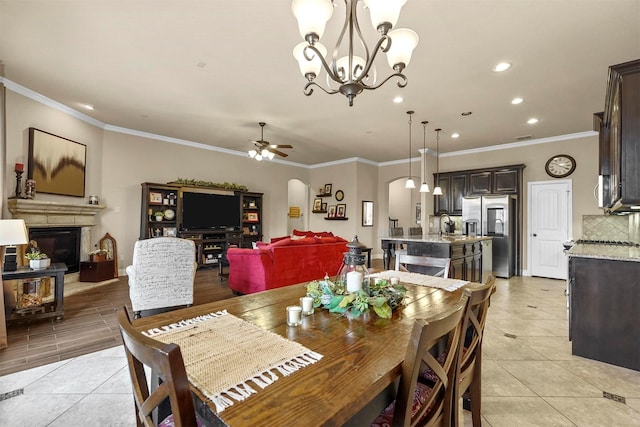 dining space with light tile patterned floors, arched walkways, a glass covered fireplace, crown molding, and ceiling fan with notable chandelier