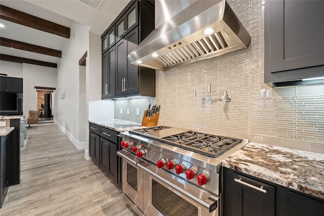 kitchen featuring decorative backsplash, glass insert cabinets, wall chimney range hood, light stone countertops, and double oven range