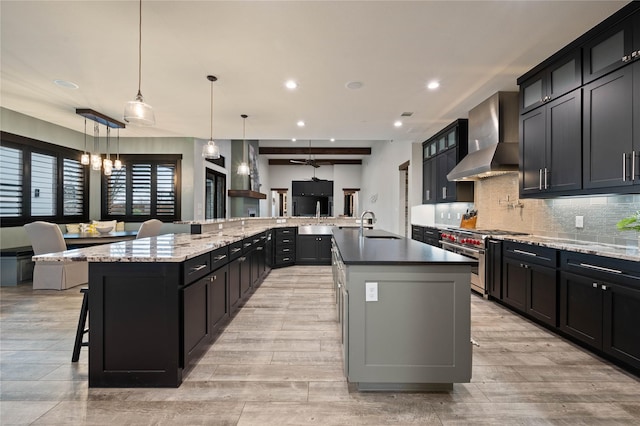 kitchen with high end stove, open floor plan, hanging light fixtures, wall chimney exhaust hood, and a large island with sink