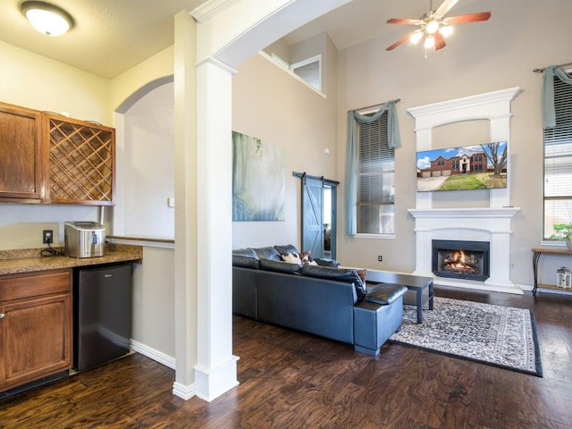 living area with dark wood-type flooring, a warm lit fireplace, a barn door, baseboards, and ceiling fan