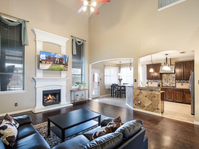 living area with baseboards, a warm lit fireplace, a high ceiling, light wood-style floors, and a ceiling fan