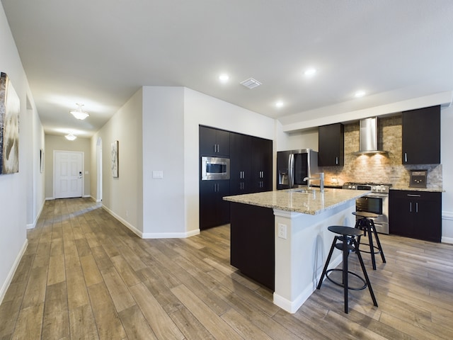 kitchen featuring an island with sink, wall chimney exhaust hood, a breakfast bar area, stainless steel appliances, and light wood-style floors