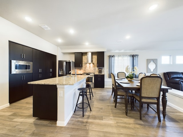 kitchen with a center island with sink, visible vents, appliances with stainless steel finishes, wall chimney range hood, and a kitchen bar