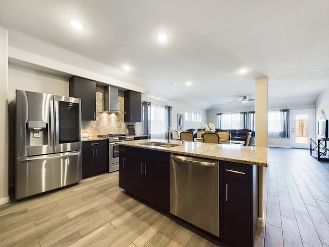 kitchen with a center island with sink, wall chimney exhaust hood, open floor plan, stainless steel appliances, and a sink