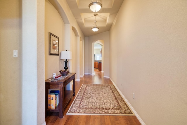 corridor with arched walkways, a raised ceiling, wood finished floors, and baseboards