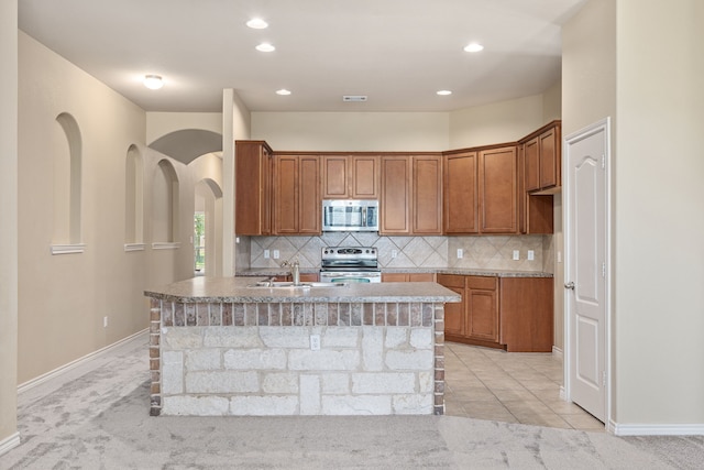 kitchen with stainless steel appliances, an island with sink, light countertops, and brown cabinetry